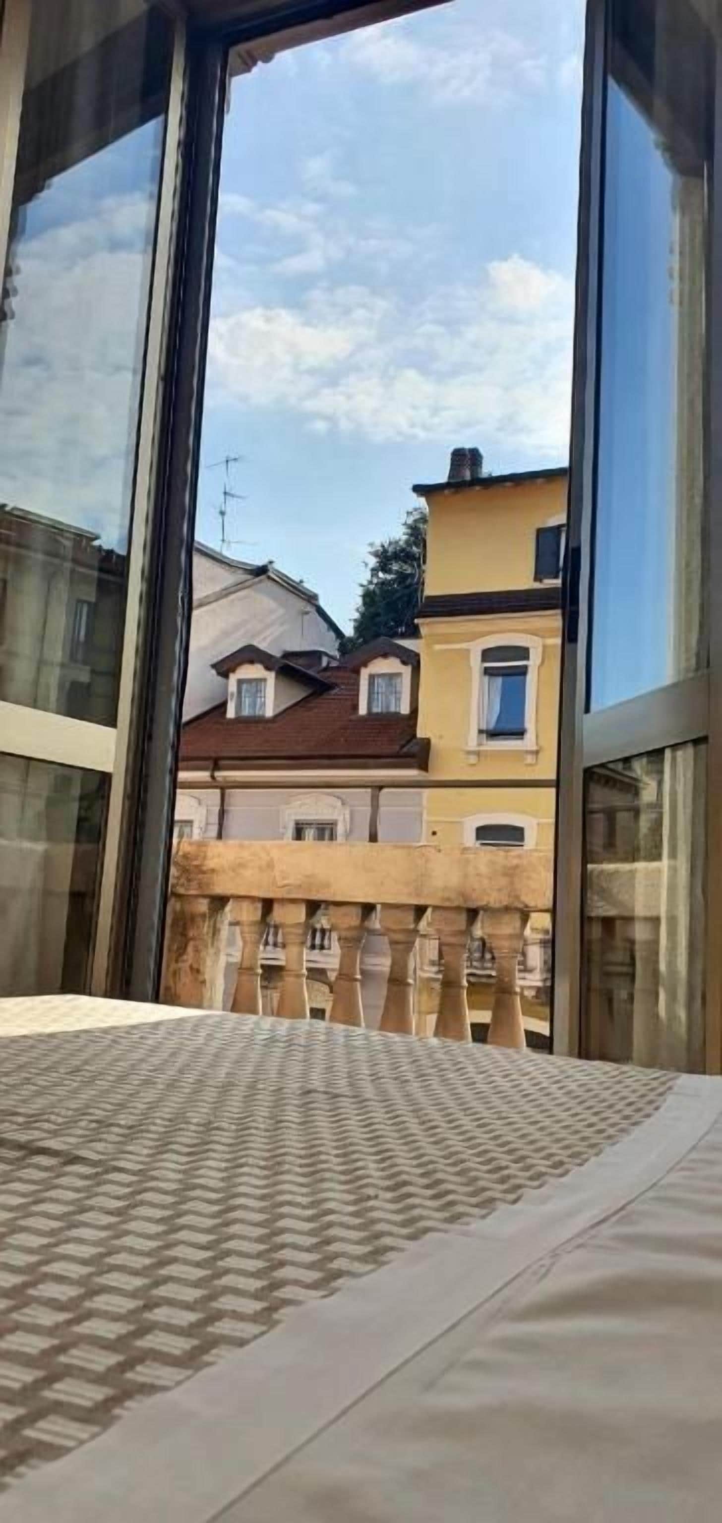 Una habitación con un ventanal grande, una cama con colcha estampada y vista a edificios cercanos.