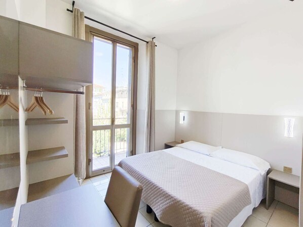 A hotel room with a bed, a desk, and a chair. There is a window with curtains, and a view of buildings outside. - Hotel Mondial (Milan)