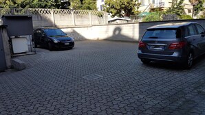A paved parking area with two cars parked, a metal fence, and a building in the background. - Hotel Mondial (Milan)