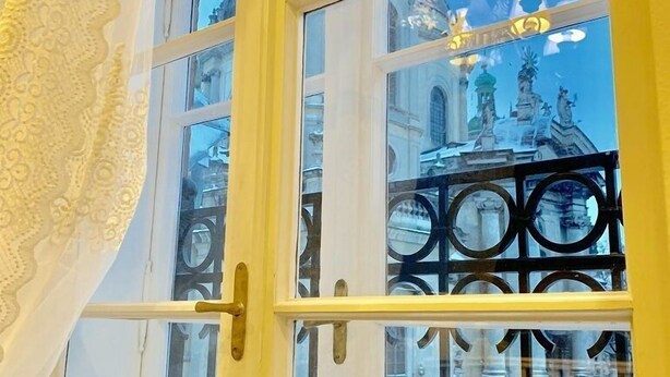 A room with a large window featuring a decorative white curtain and a view of a historic building with statues and columns. - Apartment Fodorova 1 (Lviv)