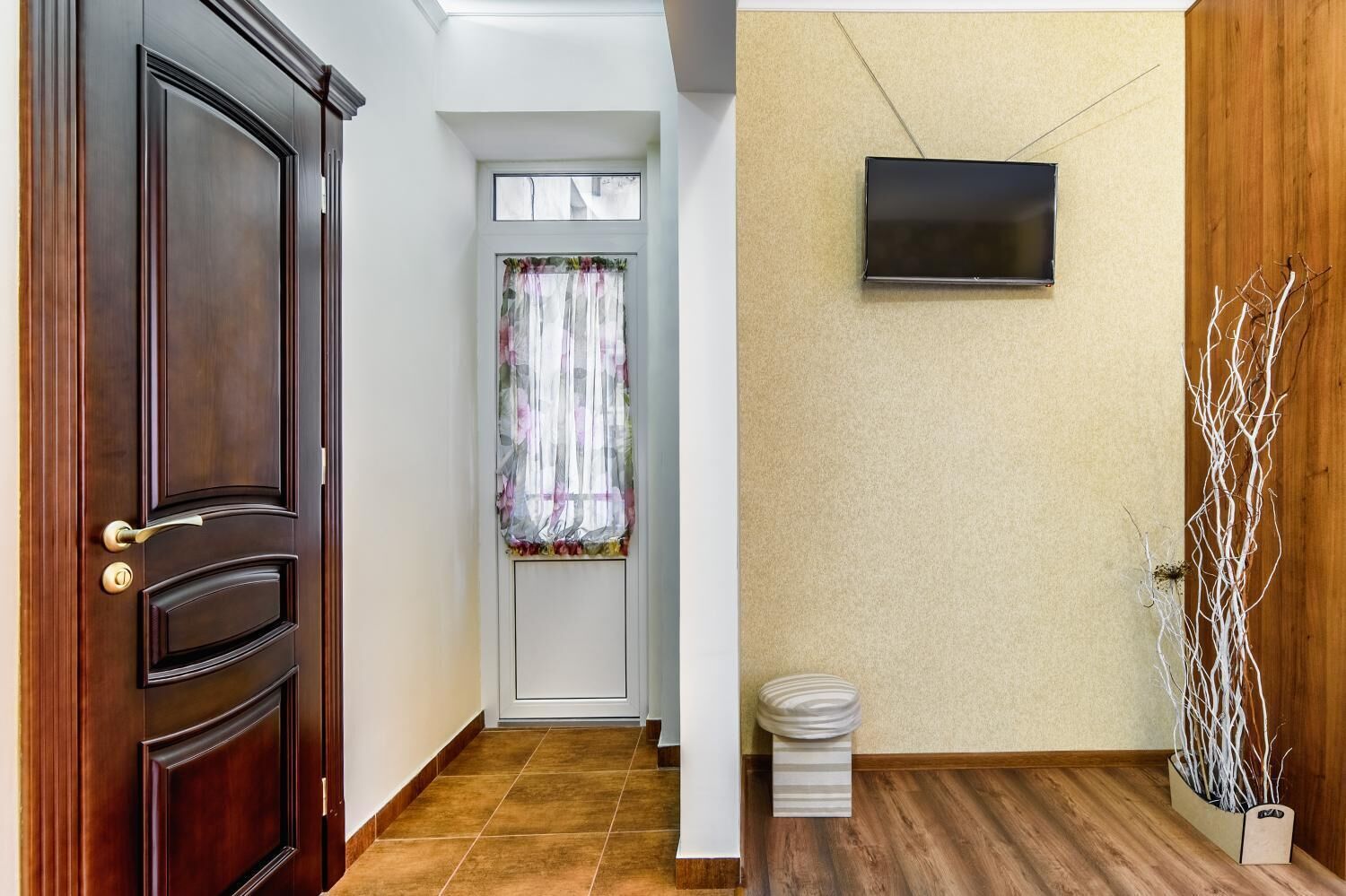A hallway with a wooden door, a window with floral curtains, a tiled floor, a small stool, and a wall-mounted TV.
