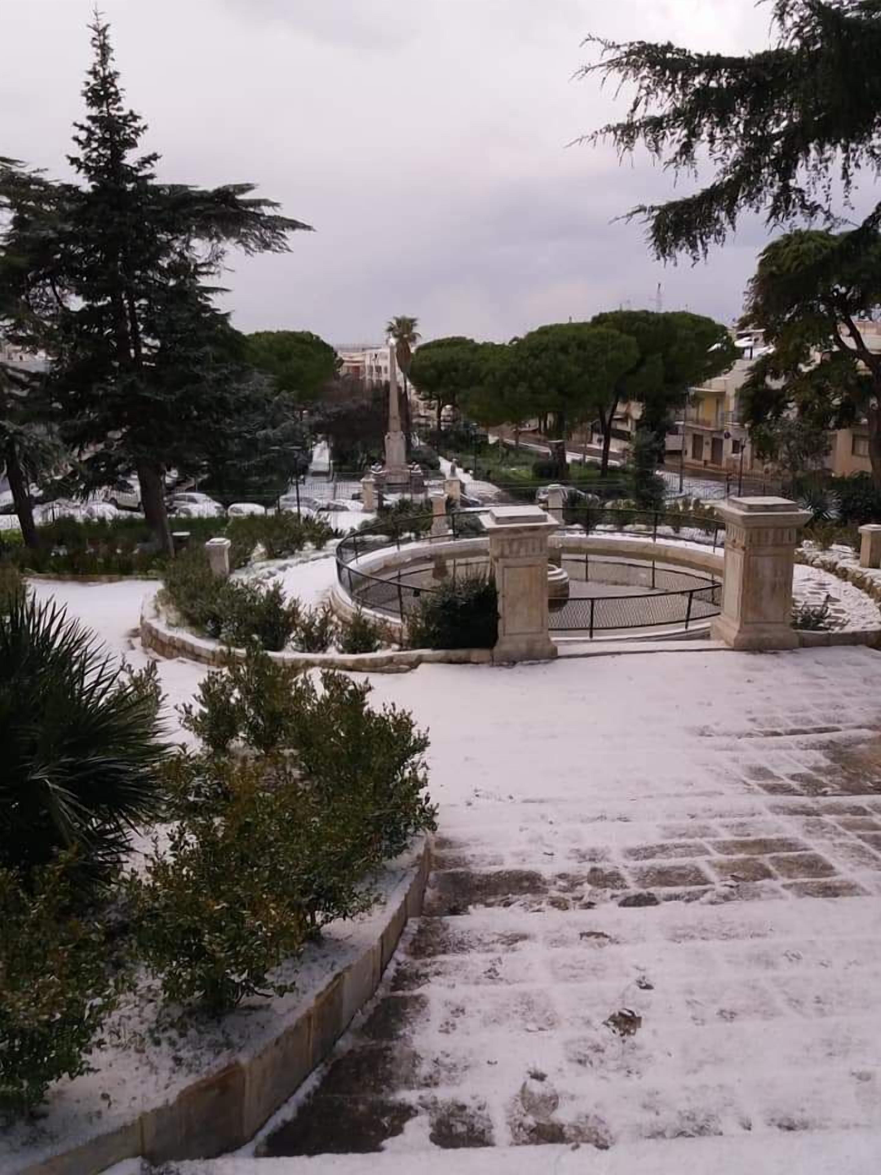 A snow-covered park with a fountain, stone benches, and trees.