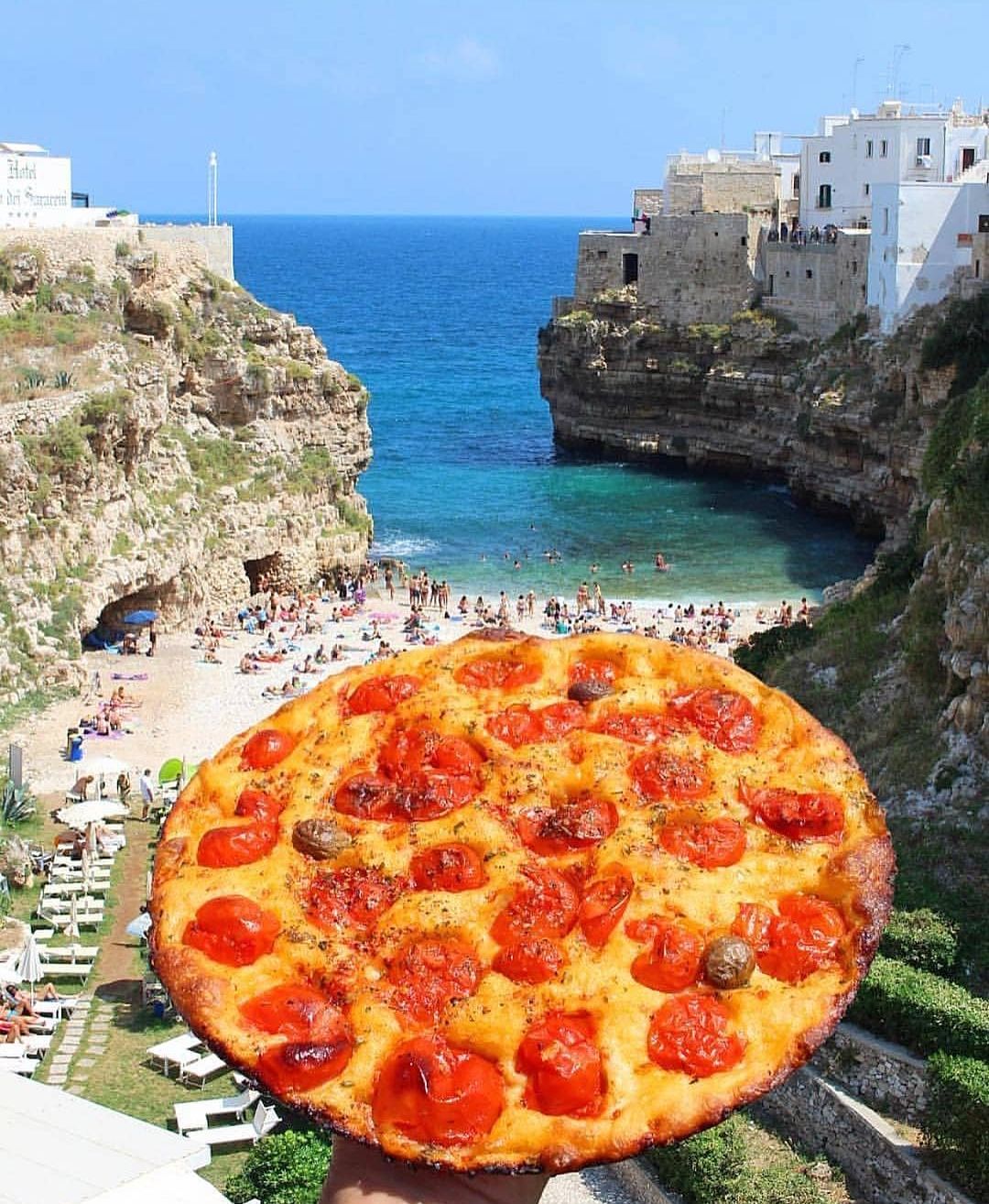 Una pizza grande con pepperoni y champiñones, sostenida frente a una vista costera con playa y edificios.