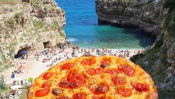 Una pizza grande con pepperoni y champiñones, sostenida frente a una vista costera con playa y edificios.