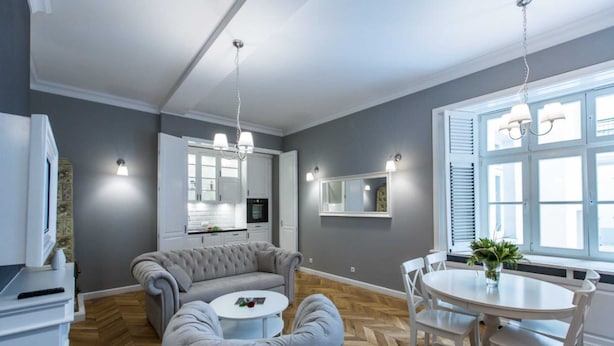 A modern kitchen with white cabinets, a grey sofa, a round dining table with chairs, and a large window with blinds.