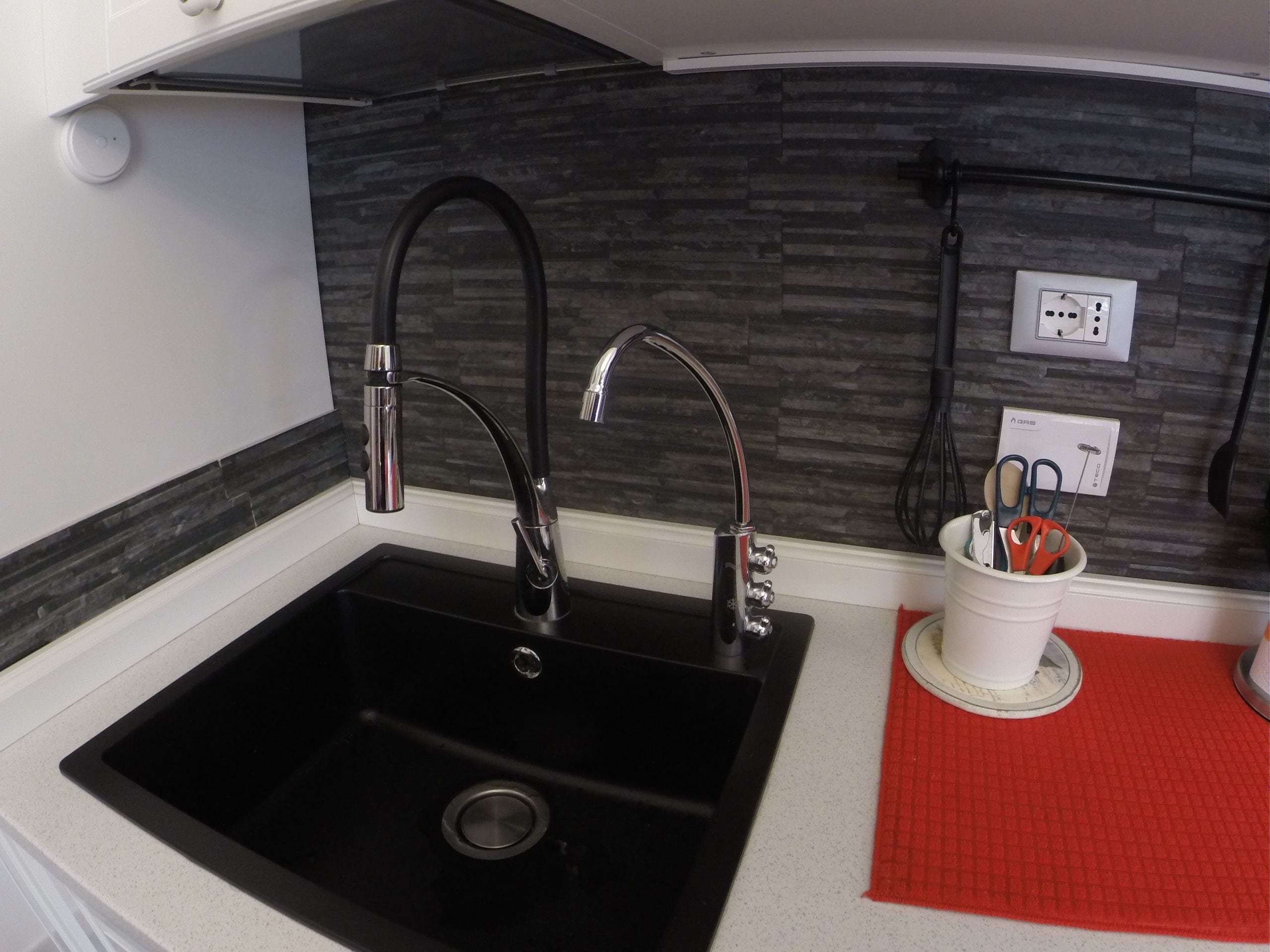 Cocina con un lavabo negro, dos grifos, un felpudo rojo y un portavasos blanco con utensilios.