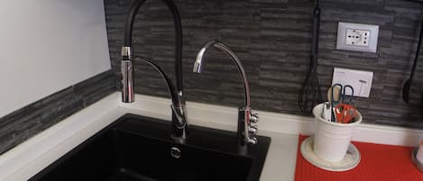 Cocina con un lavabo negro, dos grifos, un felpudo rojo y un portavasos blanco con utensilios.