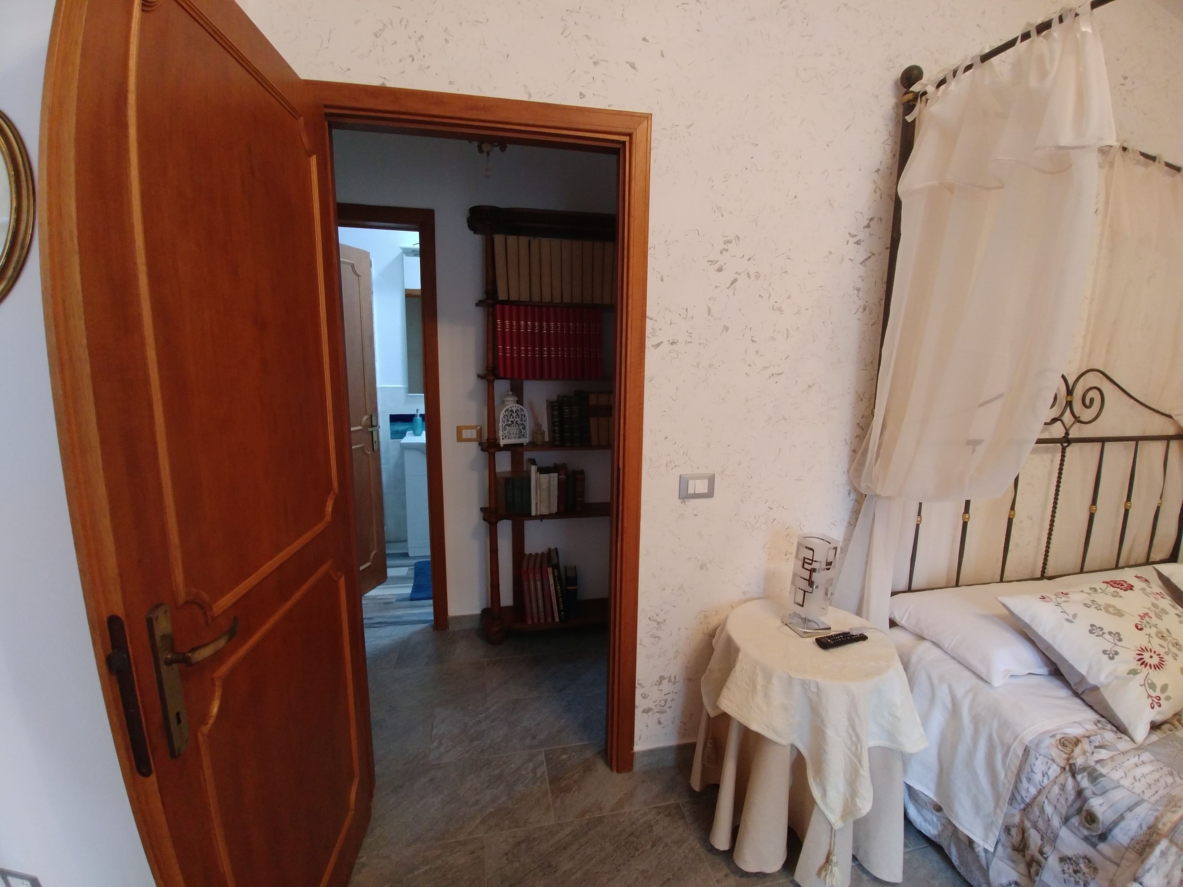 Un dormitorio con puerta de madera, mesita de noche con control remoto, cama con dosel y estantería con libros.