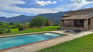 Una piscina con bordatura in pietra, un edificio in pietra con tetto in tegole e montagne sullo sfondo.