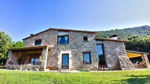 Una casa in pietra con un portico coperto, ampie finestre e vista su una collina boscosa.