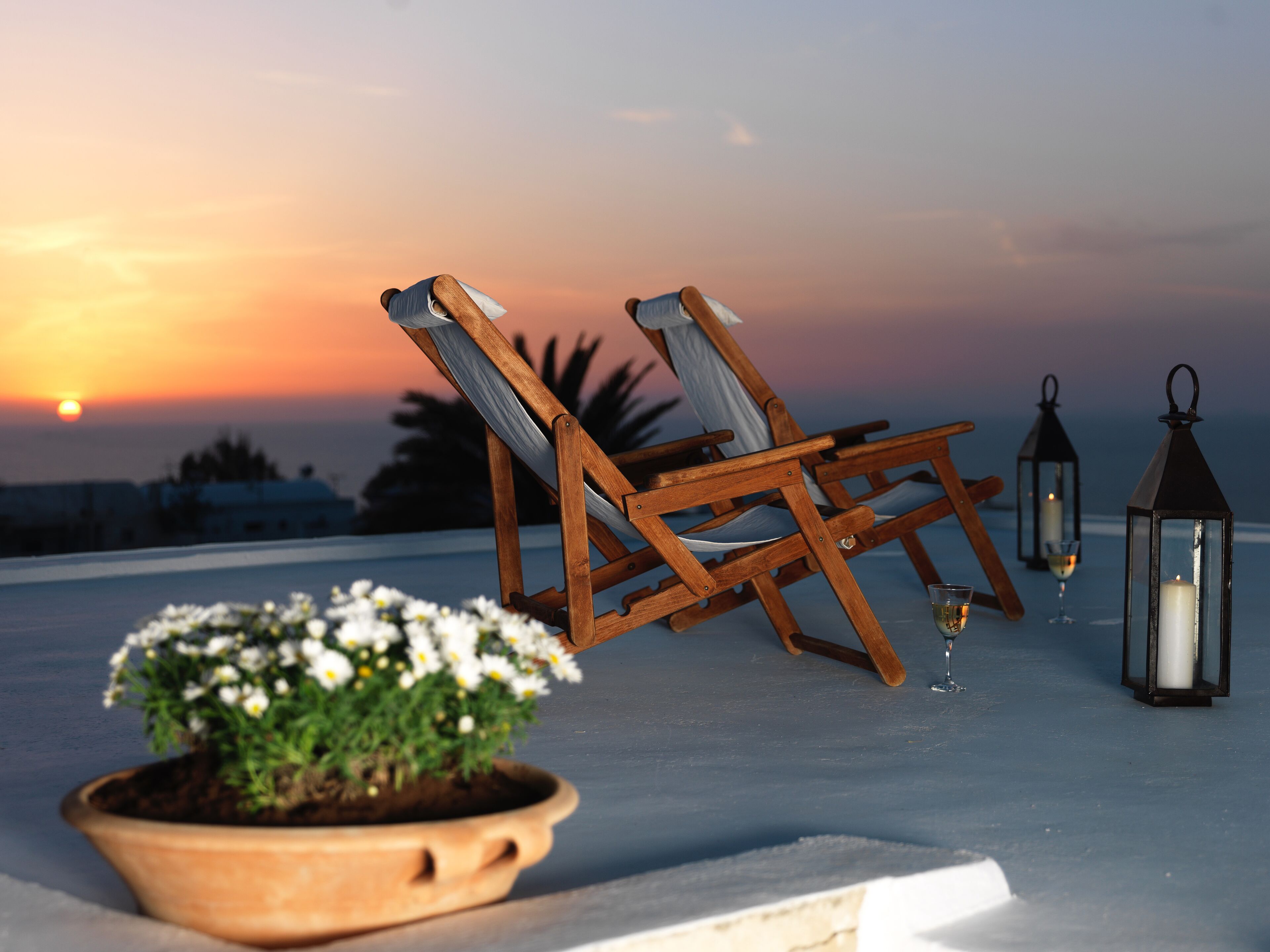 A sunset view with two wooden chairs, a potted plant, and two lanterns on a terrace.