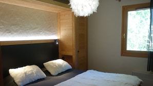 A bedroom with a bed, two white fluffy pillows, a wooden headboard, a white fluffy throw, a window with a view of greenery, and a ceiling light fixture. - 3-room apartment with balcony and garage near the slopes (La Clusaz)