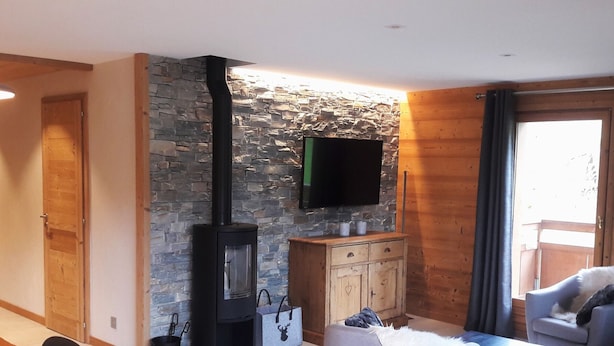 A cozy living room with a stone wall, a fireplace, a television, and a dining area. - 3-room apartment with balcony and garage near the slopes (La Clusaz)