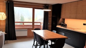 A modern kitchen with a wooden table and chairs, a built-in stove, and a view of a wooden deck through large windows. - 3-room apartment with balcony and garage near the slopes (La Clusaz)