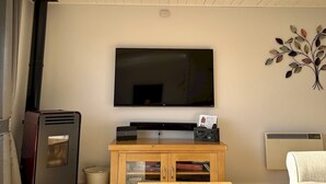 Uma sala de estar com televisão de tela plana na parede, uma mesa de centro de madeira, um sofá, uma lareira e um radiador.