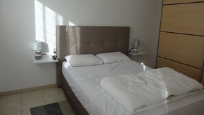 A neatly made bed with white linens and pillows, a bedside lamp, and a wall-mounted shelf with another lamp.