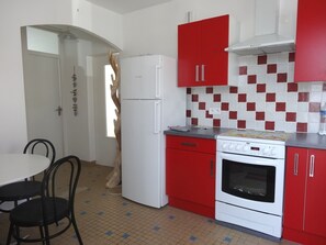 Cozinha compacta com armários vermelhos, geladeira branca e fogão. Há uma mesa de jantar com cadeiras e piso de azulejos.