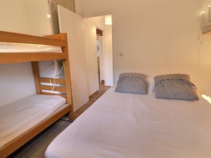 Quarto com uma cama de solteiro e uma beliche, ambas com roupa de cama branca. A cama de solteiro tem uma cabeceira de madeira e a beliche, uma parede branca.