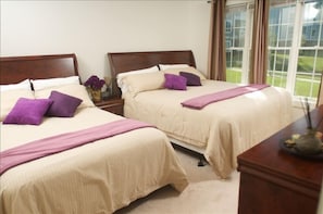 Two double beds with beige bedspreads and purple pillows, a nightstand, and a vase with flowers.