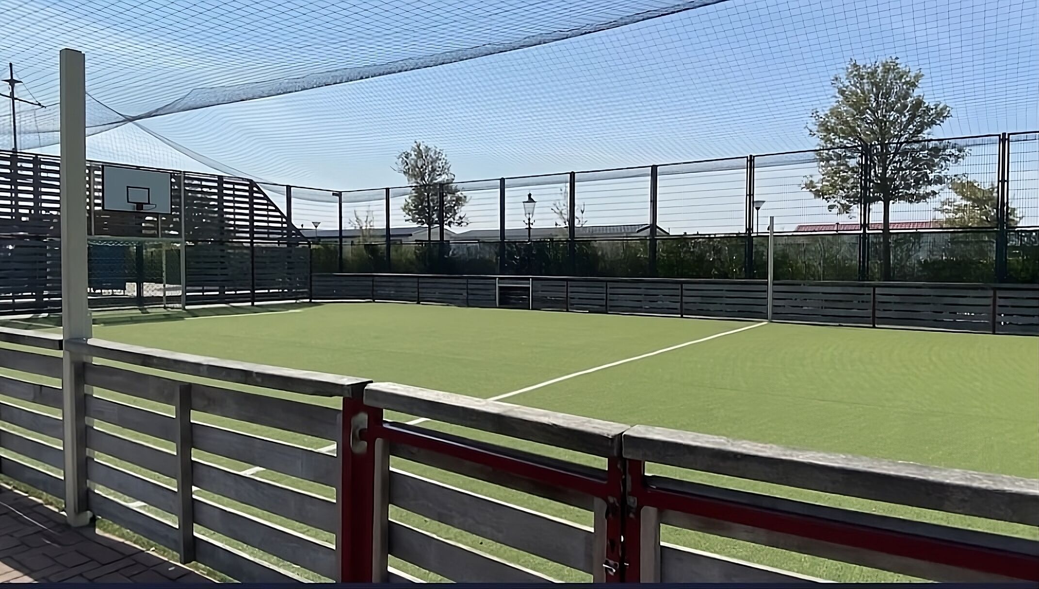 Ein Tennisplatz im Freien mit Netz, umgeben von einer Umzäunung und Zuschauerrängen.