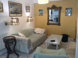 A cozy room with a bed, a small table, and a chair. There are wall-mounted lamps, a mirror, and framed pictures on the walls.