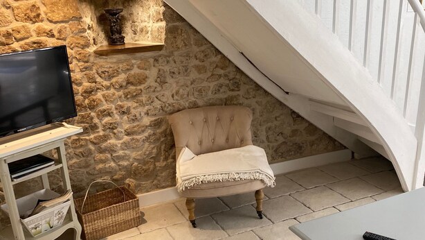 A cozy living room with a stone wall, a television on a stand, a cushioned chair, a table, and a staircase.