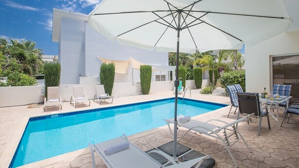 A swimming pool with lounge chairs and umbrellas, surrounded by a modern building and greenery. - Villa Molly, Lovely 3BDR Protaras Villa with Pool, Close to Fig Tree Bay Beach (Protaras)