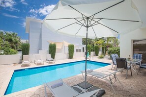 A swimming pool with lounge chairs and umbrellas, surrounded by a modern building and greenery. - Villa Molly, Lovely 3BDR Protaras Villa with Pool, Close to Fig Tree Bay Beach (Protaras)