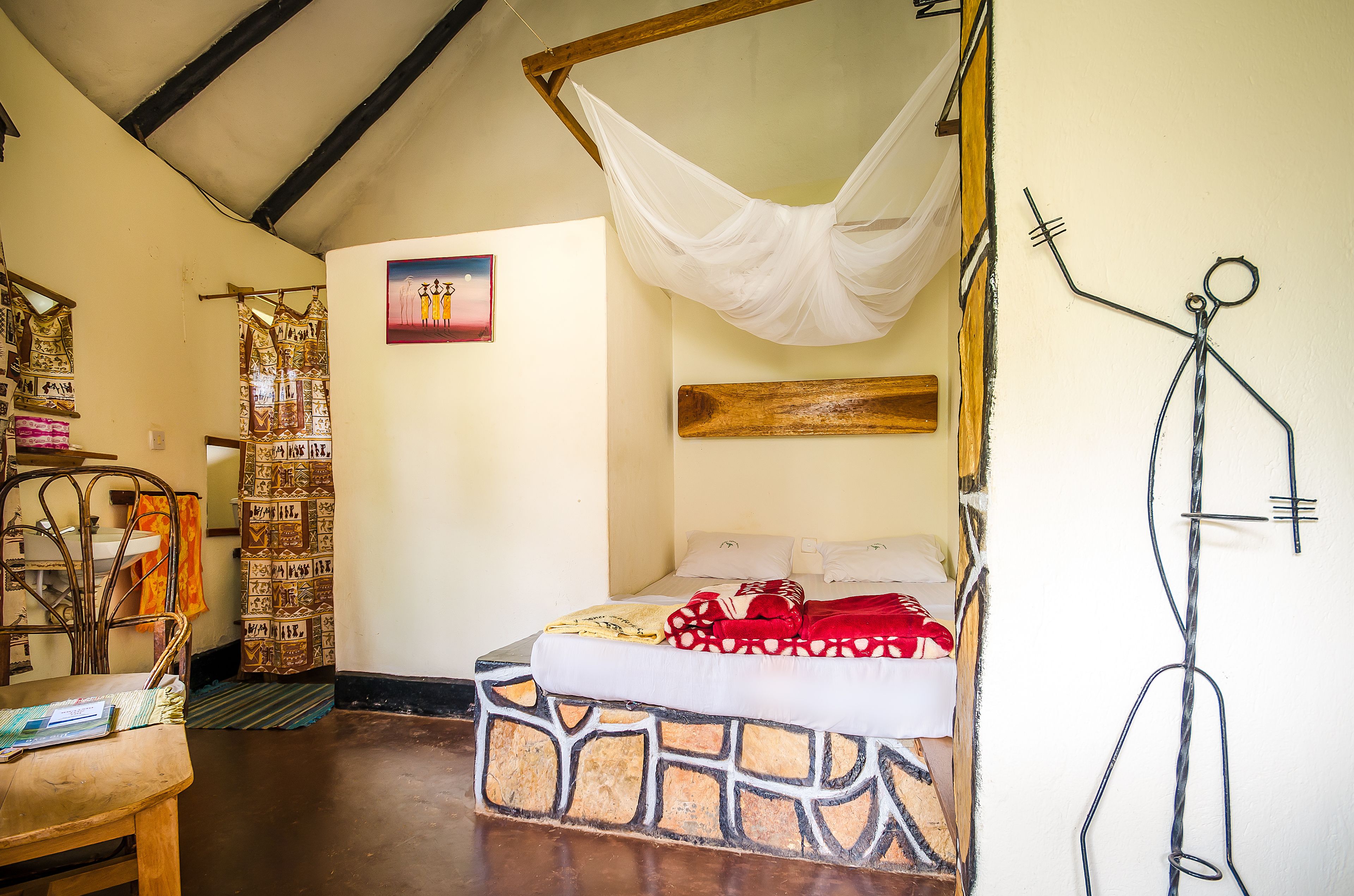 A room with a bed, a wooden bench, a wall with a picture, and a hanging sculpture.
