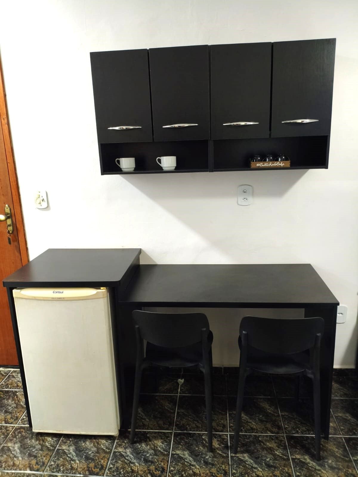 Una kitchenette pequeña con un refrigerador, una cubierta negra y un mueble superior.