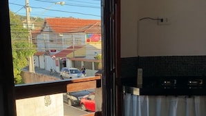 A room with a bed, a small table with a towel and flowers, a view of the street outside, and a kitchen area with a tiled backsplash. - Melhores Suites (Arraial do Cabo)