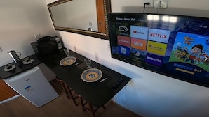 A compact hotel room with a kitchenette, a dining area with two chairs and a table, and a TV displaying various streaming services. - Melhores Suites (Arraial do Cabo)