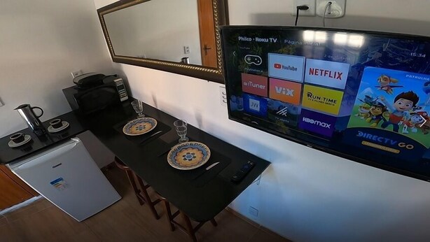 A compact hotel room with a kitchenette, a dining area with two chairs and a table, and a TV displaying various streaming services. - Melhores Suites (Arraial do Cabo)