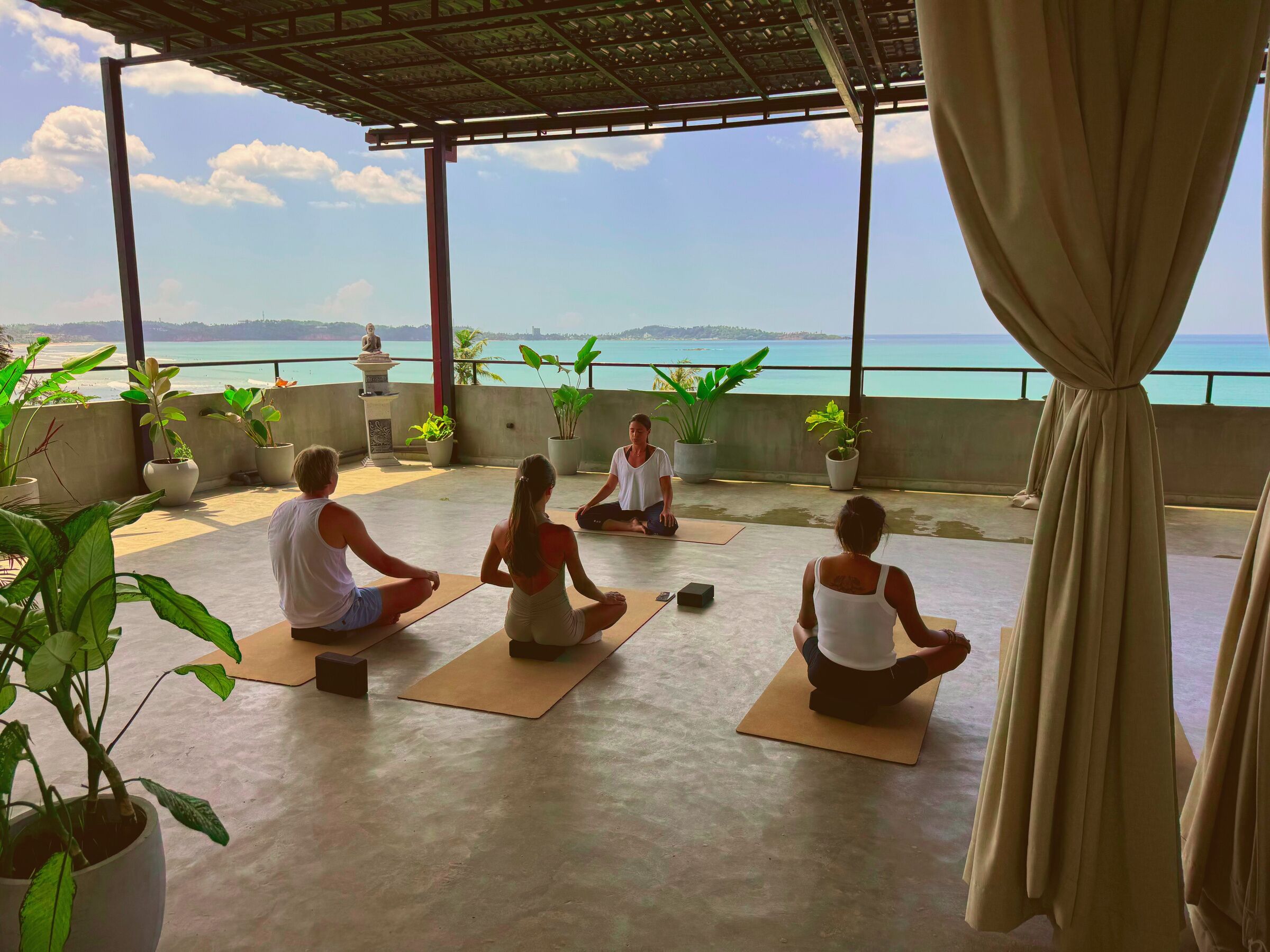 A group of people meditating on outdoor mats with potted plants and a statue, overlooking a body of water.