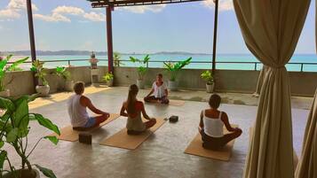 A group of people meditating on outdoor mats with potted plants and a statue, overlooking a body of water.