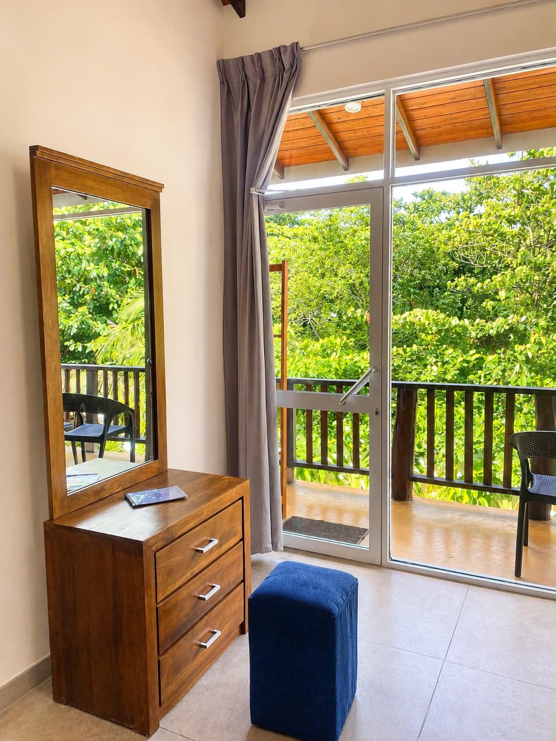 A room with a wooden dresser, a large mirror, a blue ottoman, and a sliding glass door leading to a balcony with a view of greenery.