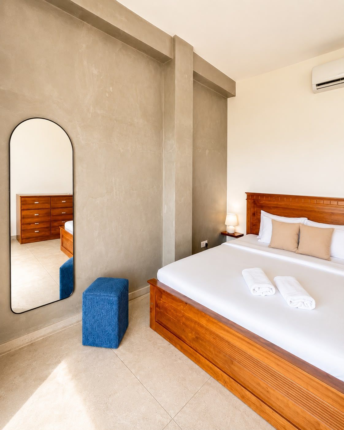 Une chambre d’hôtel avec un grand lit, une tête de lit en bois, une table de chevet avec une lampe, un miroir et un pouf bleu.