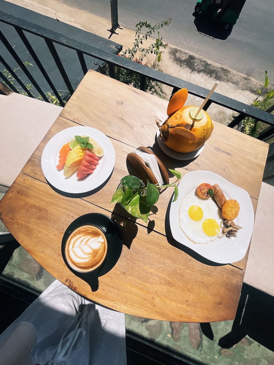 Una mesa de madera con alimentos para el desayuno, como fruta, huevos y una bebida, al aire libre con vista a la calle.