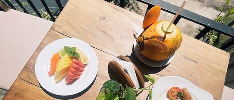 Una mesa de madera con alimentos para el desayuno, como fruta, huevos y una bebida, al aire libre con vista a la calle.