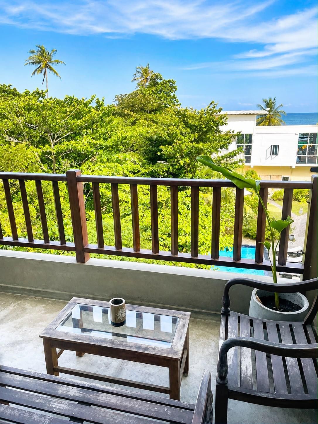 A balcony with a wooden table and chairs, a potted plant, and a view of a swimming pool and buildings.