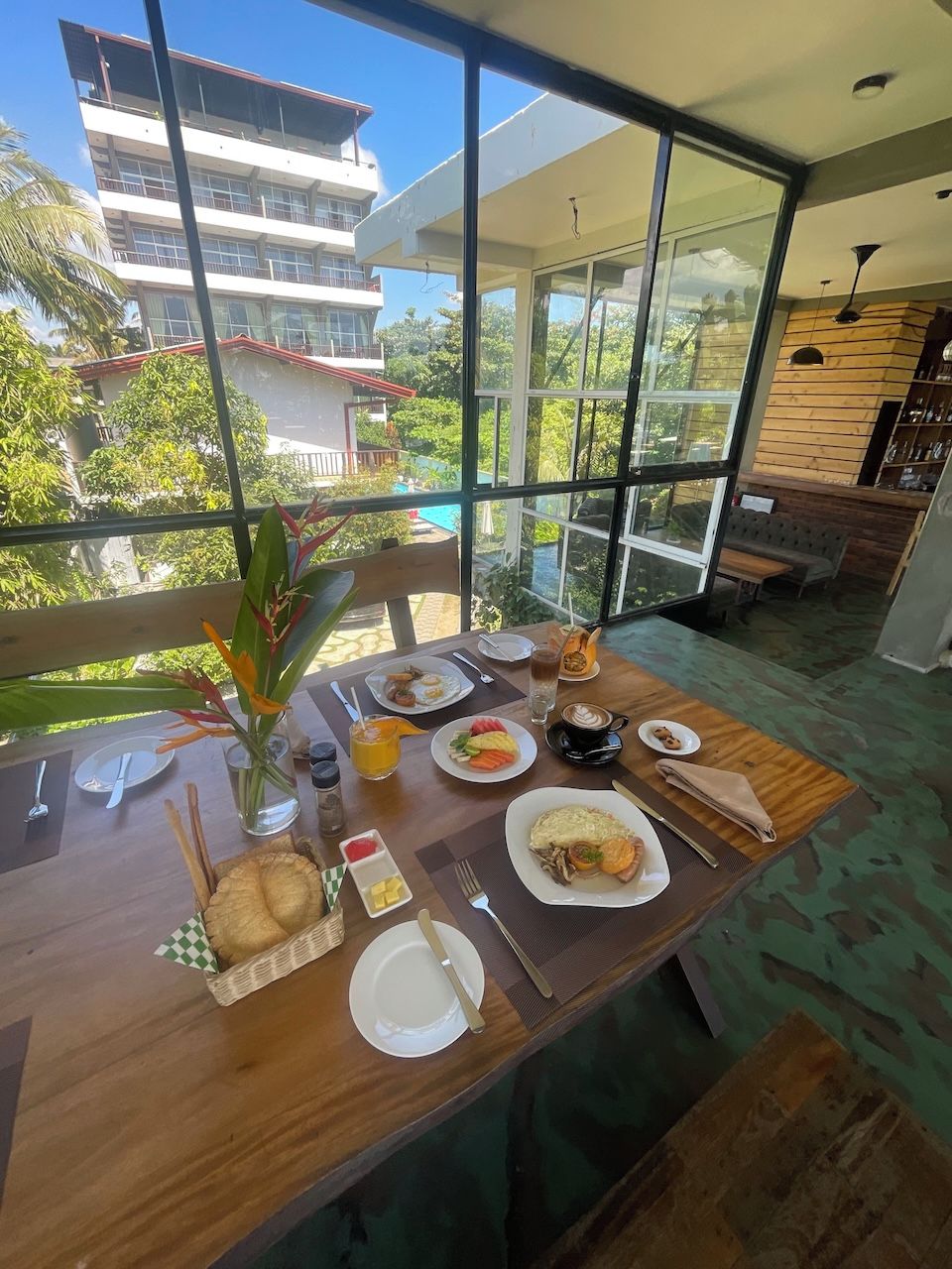 A dining table with breakfast items, a vase with flowers, and a view of a building and trees through large windows.