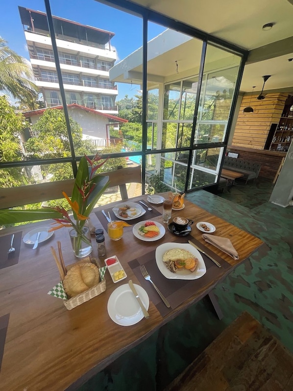 Una mesa de comedor con alimentos para el desayuno, un florero con flores y vista a un edificio y árboles a través de grandes ventanales.