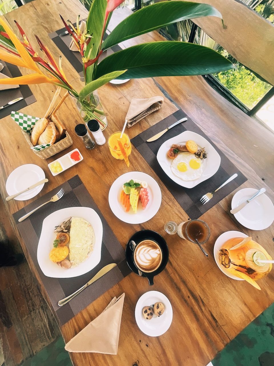 A wooden dining table with various breakfast items including eggs, toast, and fruit, set in a bright room with a view of greenery outside.