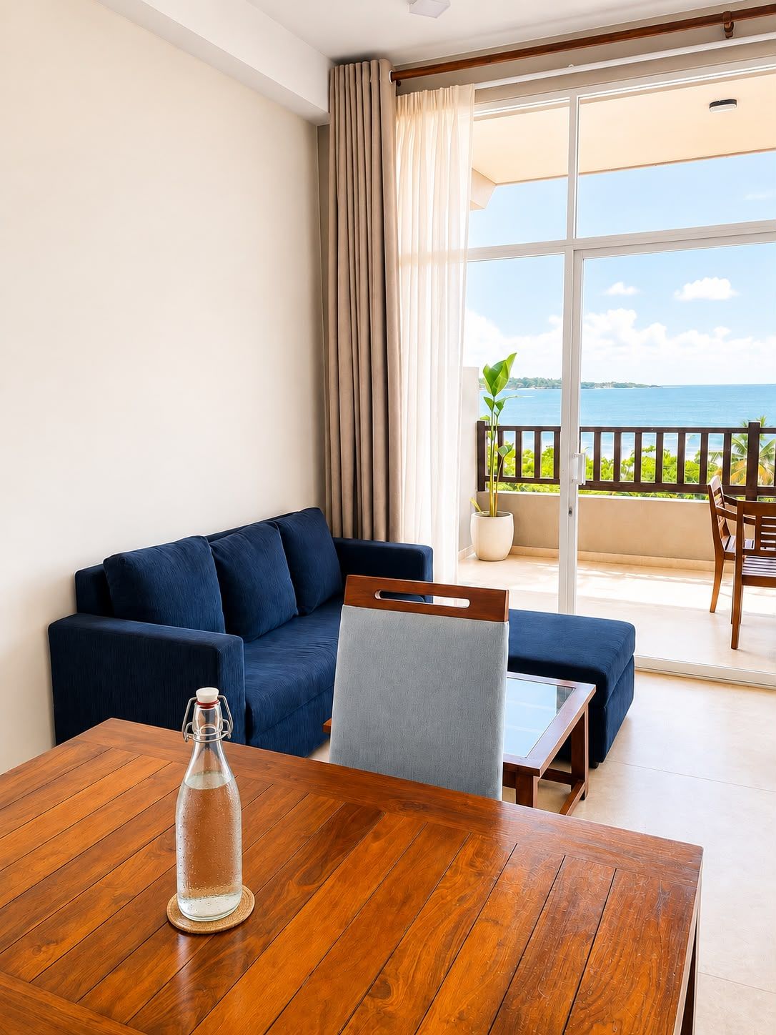 A room with a wooden table, a blue sofa, a chair, and a view of the sea through large windows.
