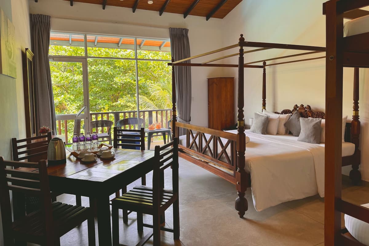 A four-poster bed with a canopy, a dining table with chairs, and a view of greenery through large windows.