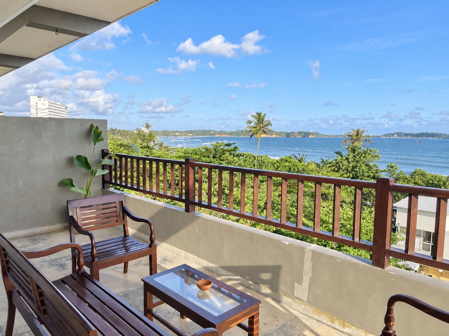 A balcony with a wooden bench, a glass-top table, and a view of the ocean and palm trees.