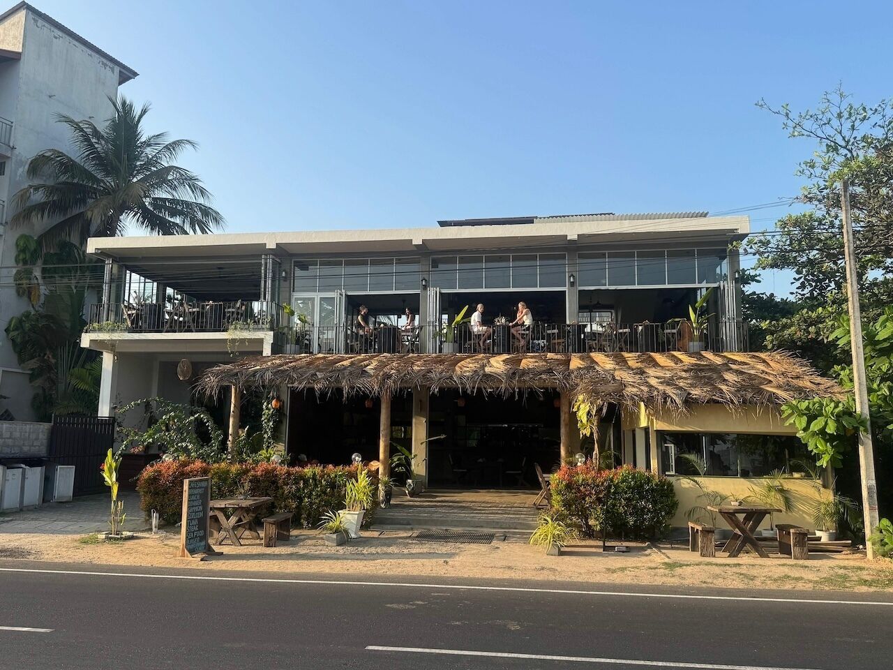 Un edificio de dos pisos con un área de estar techada al aire libre y una estructura con techo de paja.
