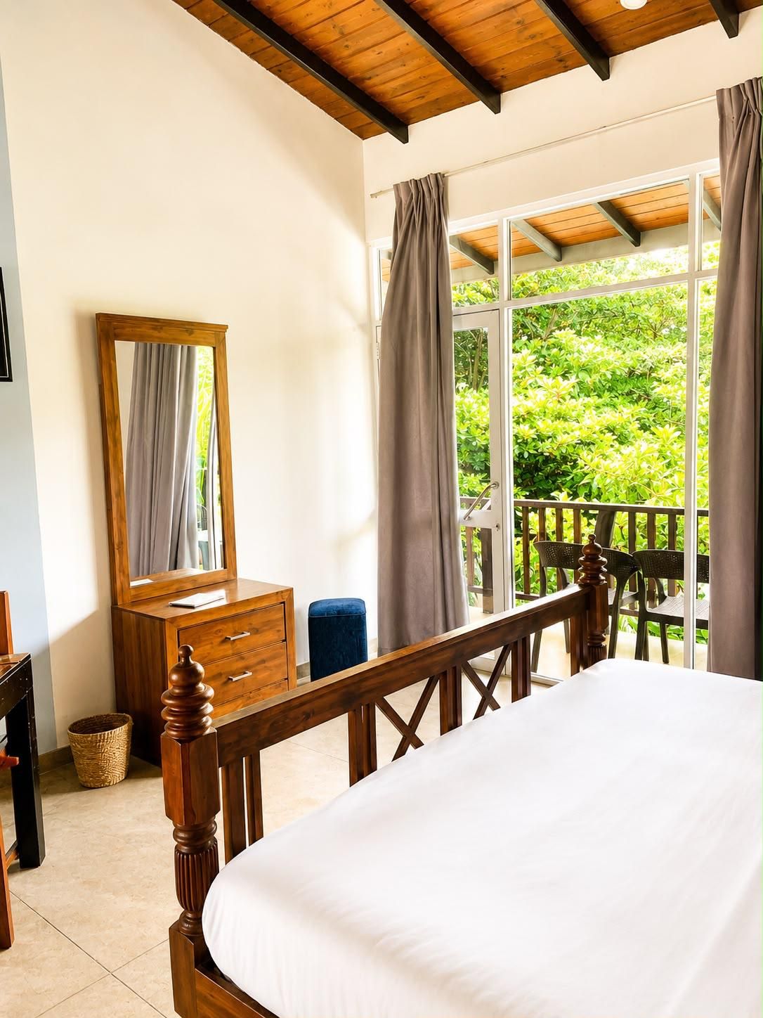 A bedroom with a wooden bed, a dresser, a mirror, and a view of greenery through large windows.