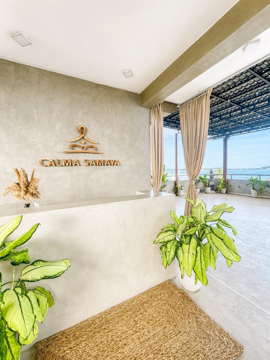 A reception area with a sign reading 'CALMA SAMAYA' and a view of the sea through large windows.
