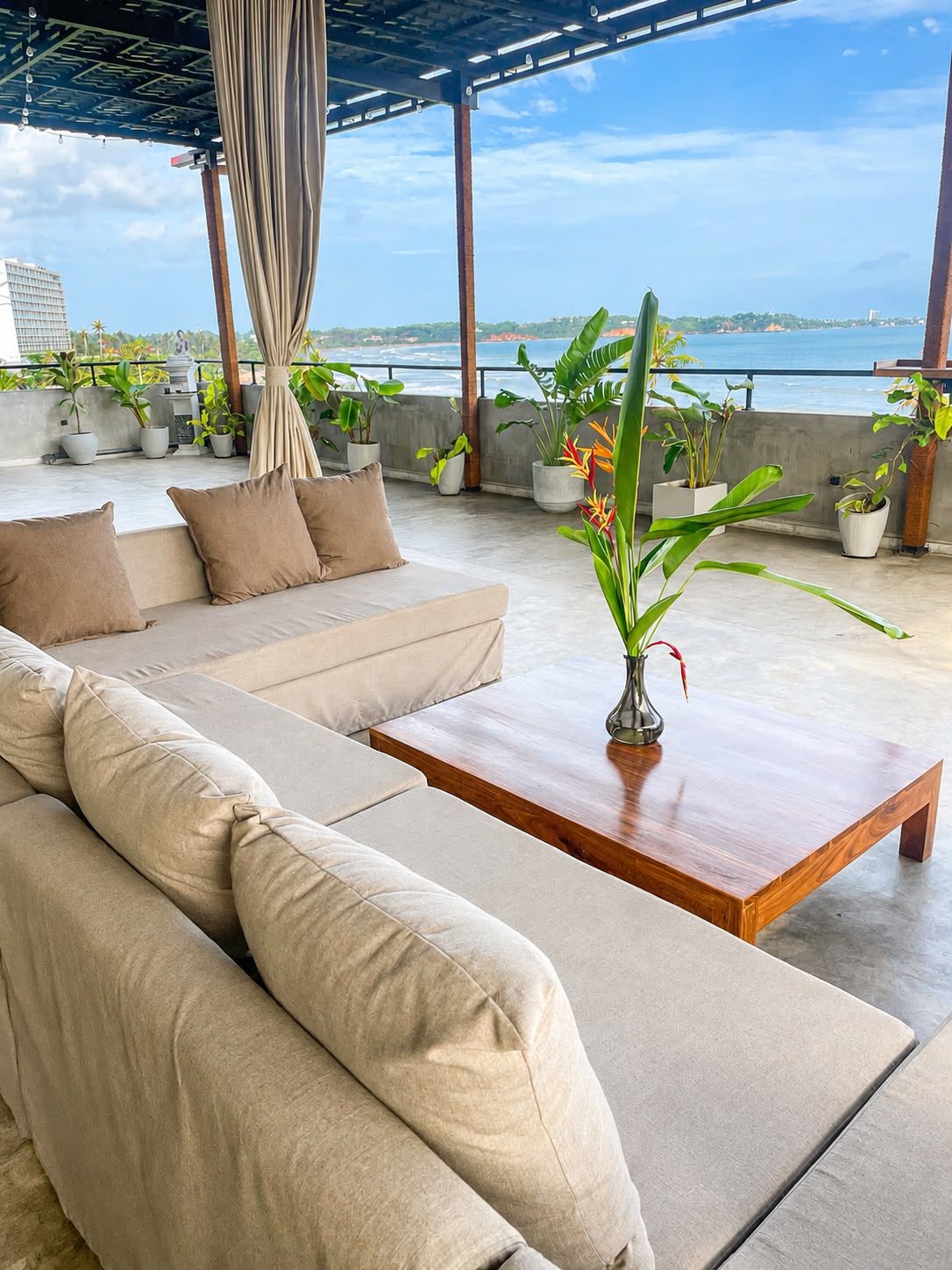 A modern outdoor seating area with a sofa and a wooden coffee table, surrounded by potted plants and overlooking a body of water.